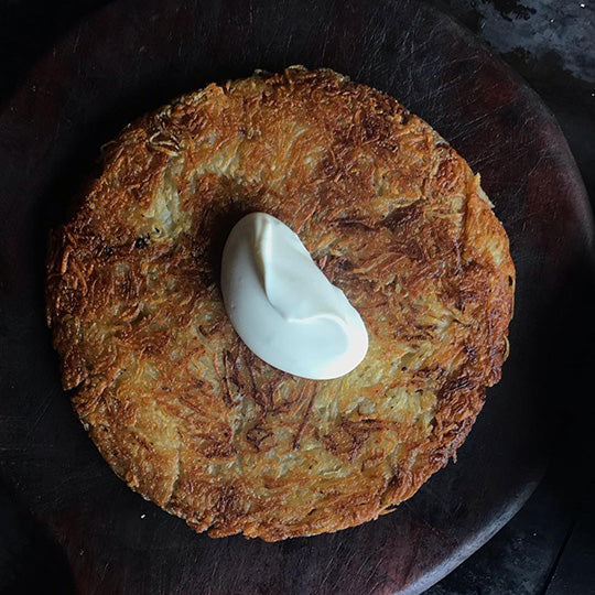 A round golden-brown hash brown or potato pancake topped with a dollop of white sour cream, presented on a dark wooden plate.