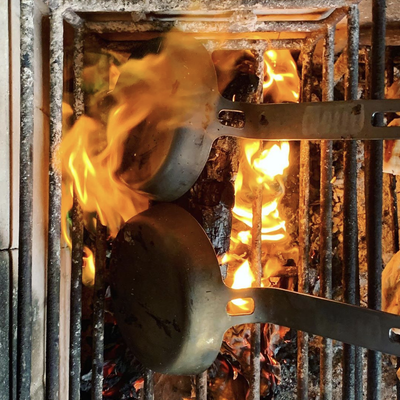 Two DARTO dark carbon steel skillets with metal handles resting on a black metal grill grate over an active fire with bright orange flames.