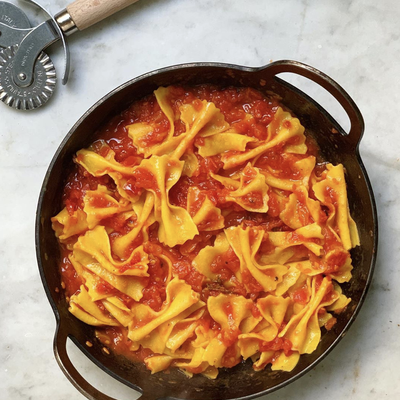 A top-down view of a rustic dark DARTO carbon steel Paella filled with bowtie pasta in a rich tomato sauce, set on a light marble surface with a metal pizza cutter visible in the upper left corner.
