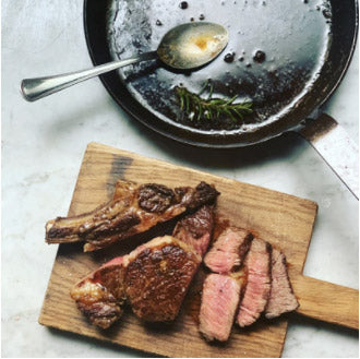 Sliced and boneless cooked steak pieces arranged on a wooden cutting board, with a dark DARTO carbon steel skillet and spoon containing rendered fat in the background on a light stone surface.