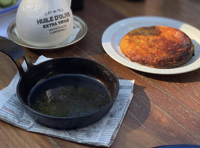 A DARTO carbon steel pan filled with dark olive oil sits on a newspaper pad on a wooden table, next to a white plate with a toasted round bread or pita and a white bottle labeled HUILE D'OLIVE EXTRA VINE.