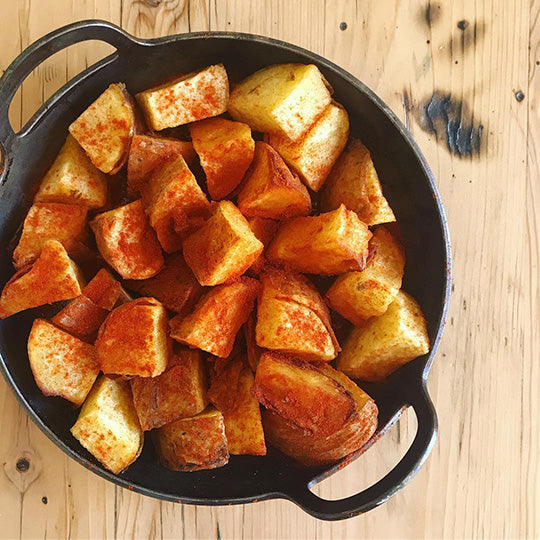 Seasoned cubed patatas bravas in a black DARTO CARBON STEEL skillet, photographed from above on a light wood tabletop.