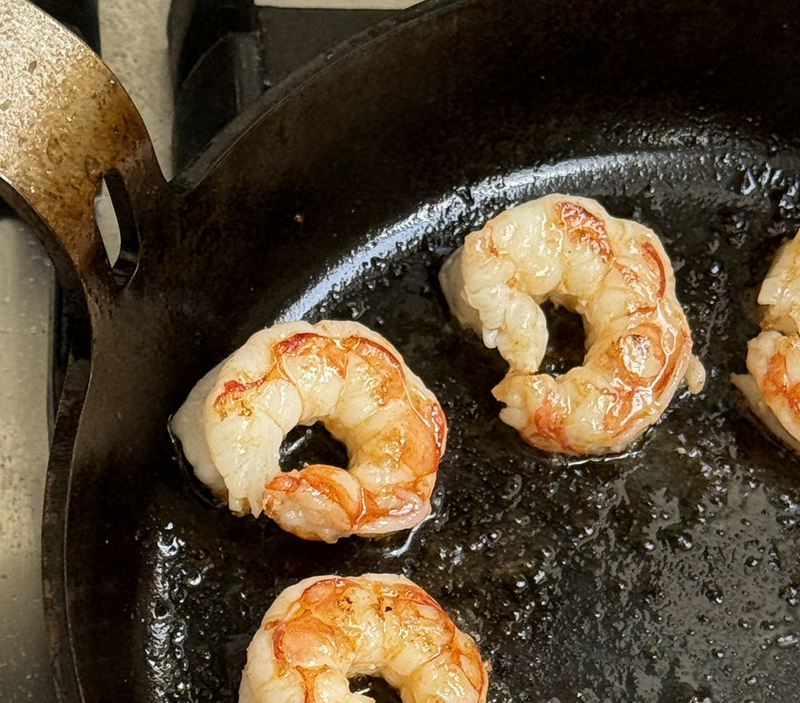 Four shrimp cooking in a black carbon steel DARTO skillet on a stovetop.